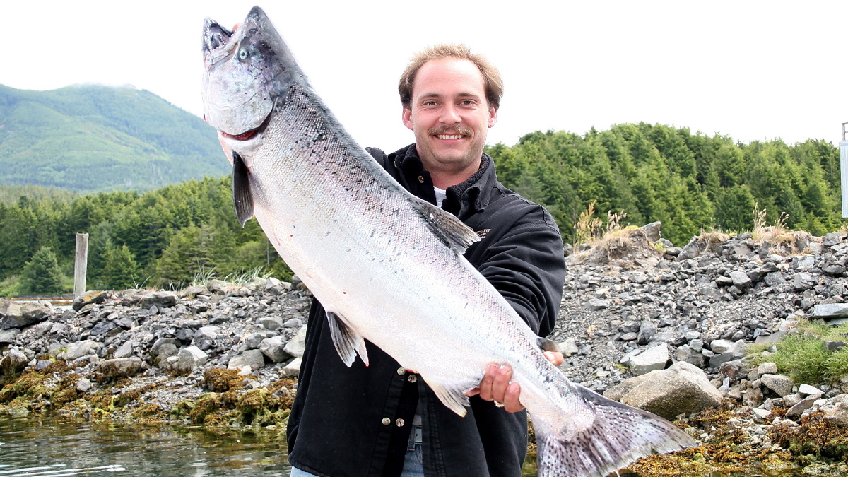 Man in a black shirt holding a huge chinook salmon in shallow water on a sunny day.
