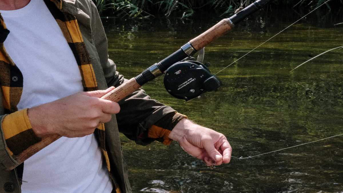 Man with a big baitcasting rod holding the line between two fingers in a stream.