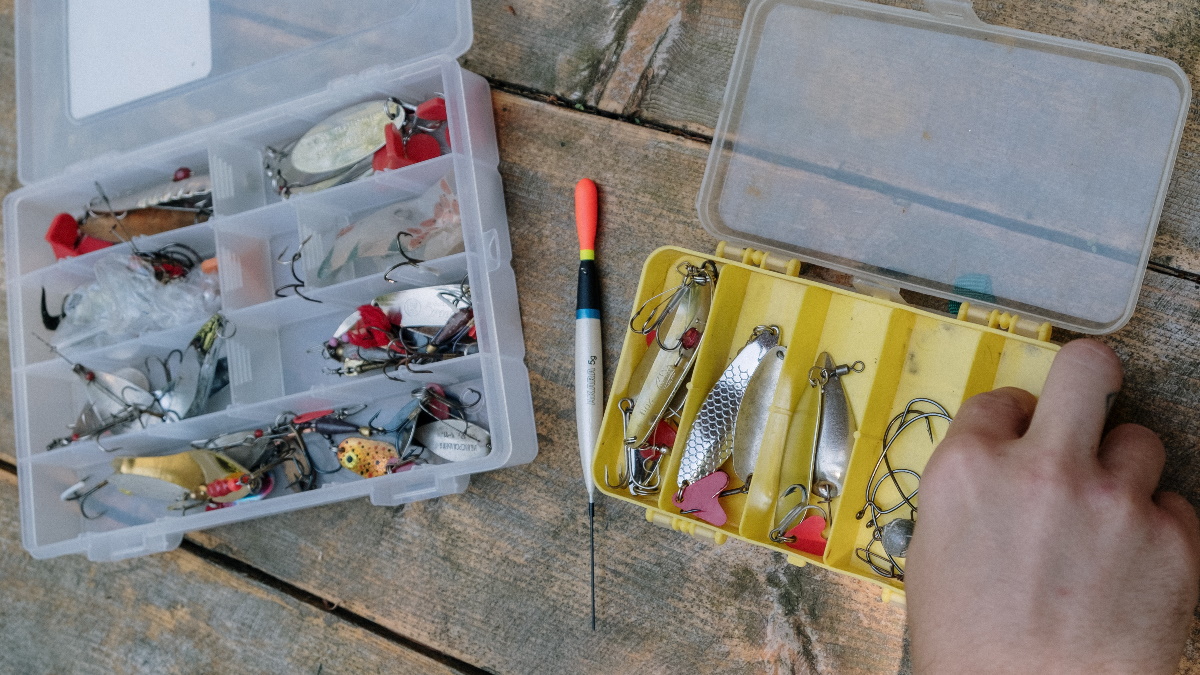 Man's hand grabbing a hook out of a tackle box next to another tackle box with spoons and a bobber on a wooden table.